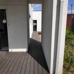 Arrur Musiał - Nowoczesny taras z desek kompozytowych w odcieniach brązu, prowadzący wzdłuż białej ściany domu z widocznym systemem rynnowym i elementami zieleni w tle.