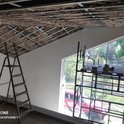 Arrur Musiał - Montaż izolacji wełną mineralną w metalowym stelażu sufitu podwieszanego, widoczne drabiny i rusztowanie przy oknie.