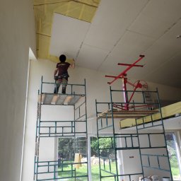 Arrur Musiał - Pracownik na rusztowaniu montuje płytę na suficie poddasza, widoczna izolacja termiczna, podnośnik do płyt i worki z materiałami budowlanymi na podłodze.