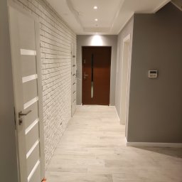 Arrur Musiał - Korytarz po remoncie z podłogą z paneli imitujących drewno, ściana wykończona białą cegłą, szara ściana z panelem alarmowym i brązowe drzwi wejściowe.