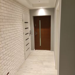 Arrur Musiał - Korytarz z białą cegłą na ścianie, drewnianymi drzwiami wejściowymi i jasnymi płytkami podłogowymi, częściowo otwarte białe drzwi po lewej stronie.