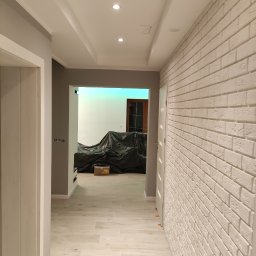 Arrur Musiał - Korytarz w trakcie remontu z wykończoną ścianą z białej cegły, podłoga wyłożona płytkami imitującymi drewno, meble zakryte folią ochronną, widoczny karton.