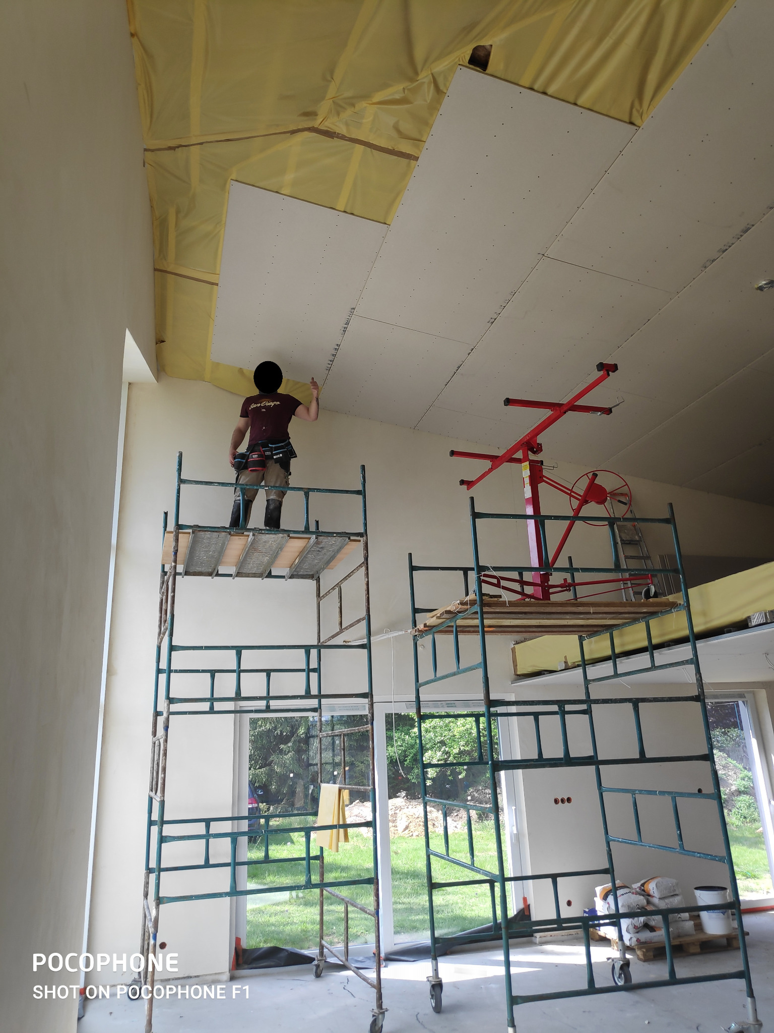 Pracownik na rusztowaniu montuje płytę na suficie poddasza, widoczna izolacja termiczna, podnośnik do płyt i worki z materiałami budowlanymi na podłodze.