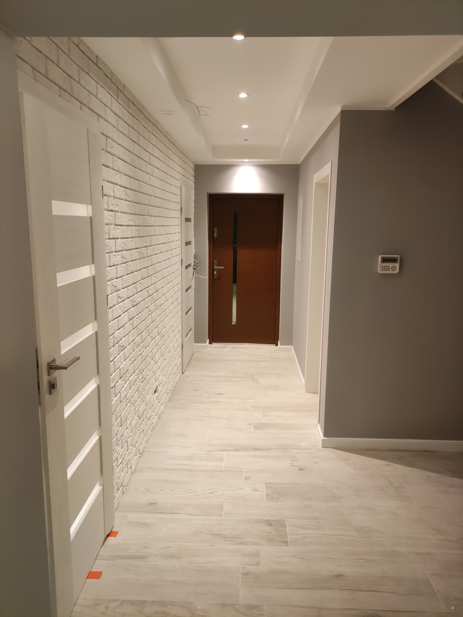 Korytarz po remoncie z podłogą z paneli imitujących drewno, ściana wykończona białą cegłą, szara ściana z panelem alarmowym i brązowe drzwi wejściowe.