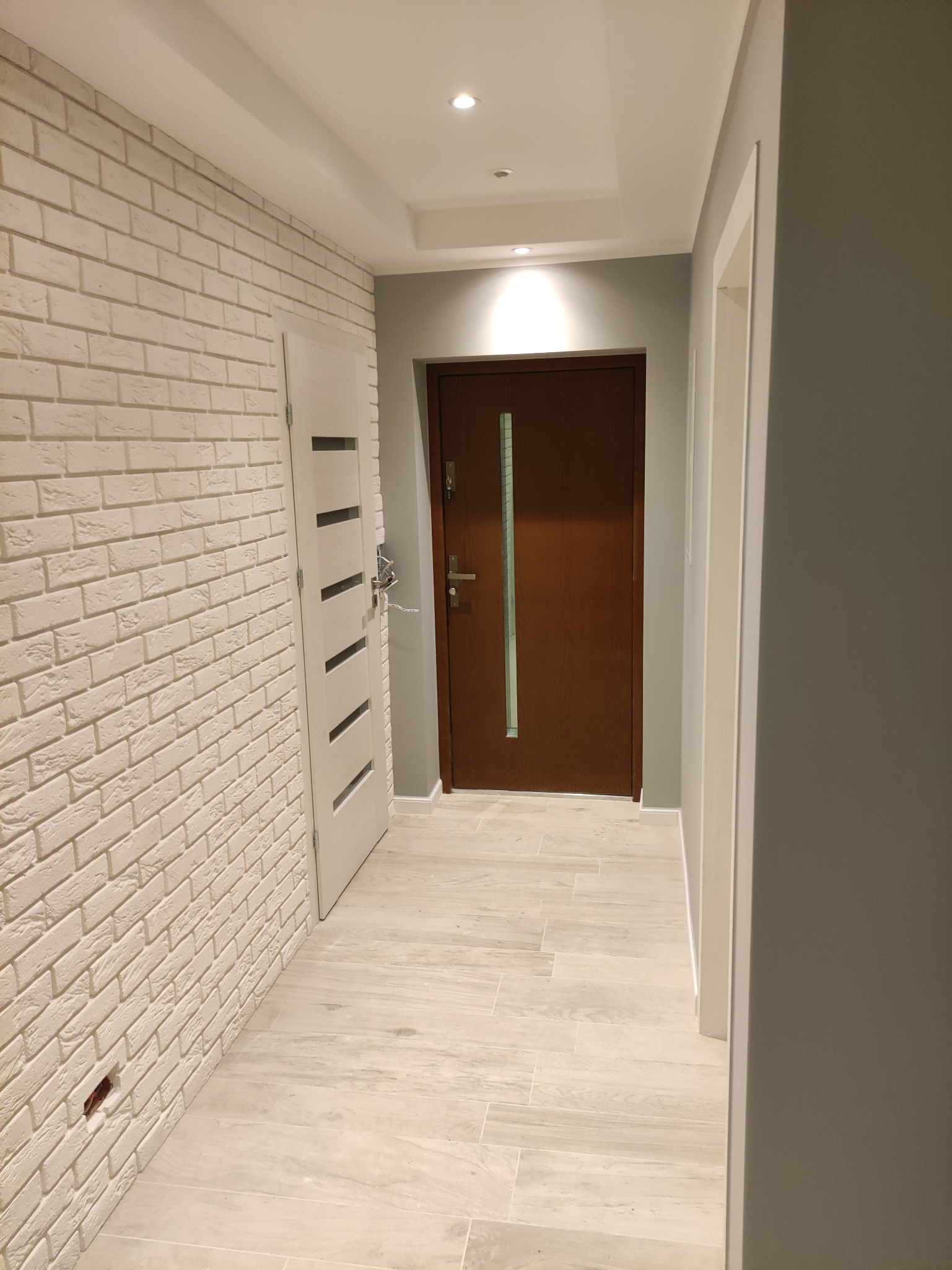Korytarz z białą cegłą na ścianie, drewnianymi drzwiami wejściowymi i jasnymi płytkami podłogowymi, częściowo otwarte białe drzwi po lewej stronie.