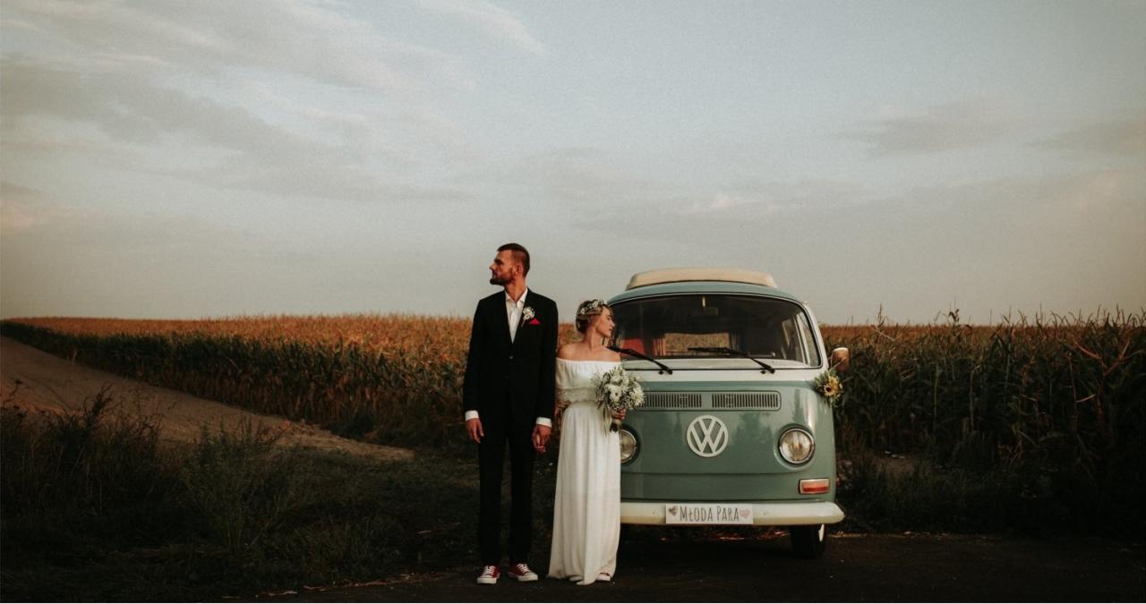 Para młoda w eleganckich strojach ślubnych stoi przed turkusowym Volkswagenem Transporterem z napisem 'Młoda Para', na tle pola kukurydzy o zachodzie słońca.