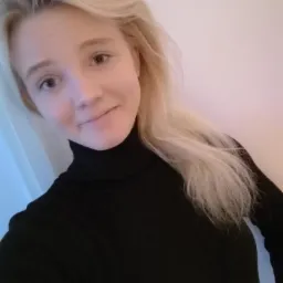 Portret uśmiechniętej blondynki w czarnym golfie na jasnym tle, z delikatnym oświetleniem.