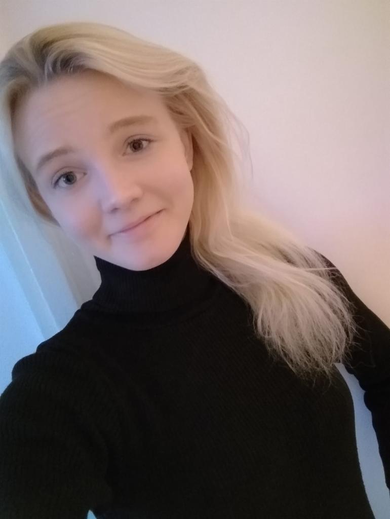 Portret uśmiechniętej blondynki w czarnym golfie na jasnym tle, z delikatnym oświetleniem.