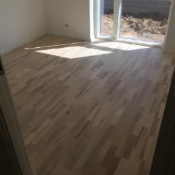 Jasne, świeżo ułożone deski parkietowe w pustym pokoju z dużym oknem, przez które wpada światło słoneczne, widoczny fragment progu drzwi.