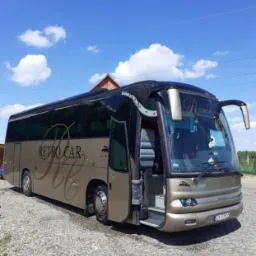Złoty autokar turystyczny z otwartymi drzwiami, zaparkowany na żwirowym placu, z napisem 'RETRO CAR' na boku i widocznym fragmentem dachu budynku w tle.