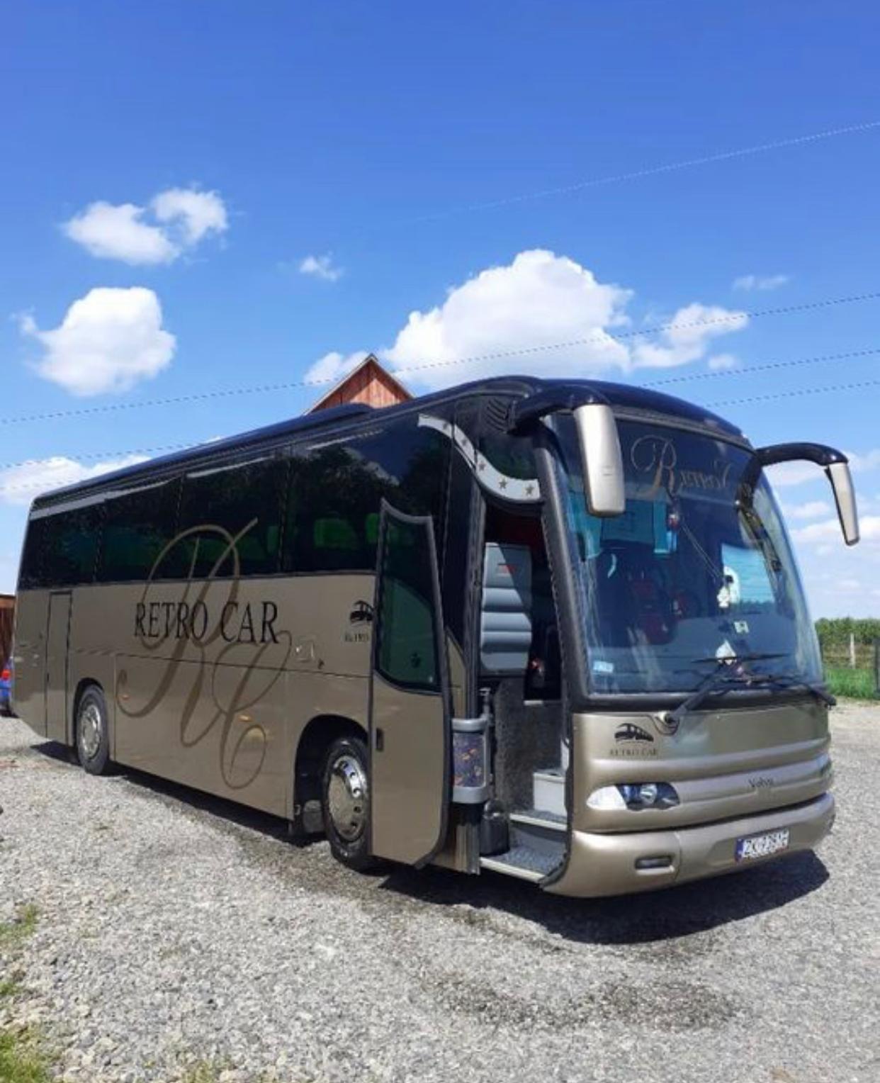 Złoty autokar turystyczny z otwartymi drzwiami, zaparkowany na żwirowym placu, z napisem 'RETRO CAR' na boku i widocznym fragmentem dachu budynku w tle.