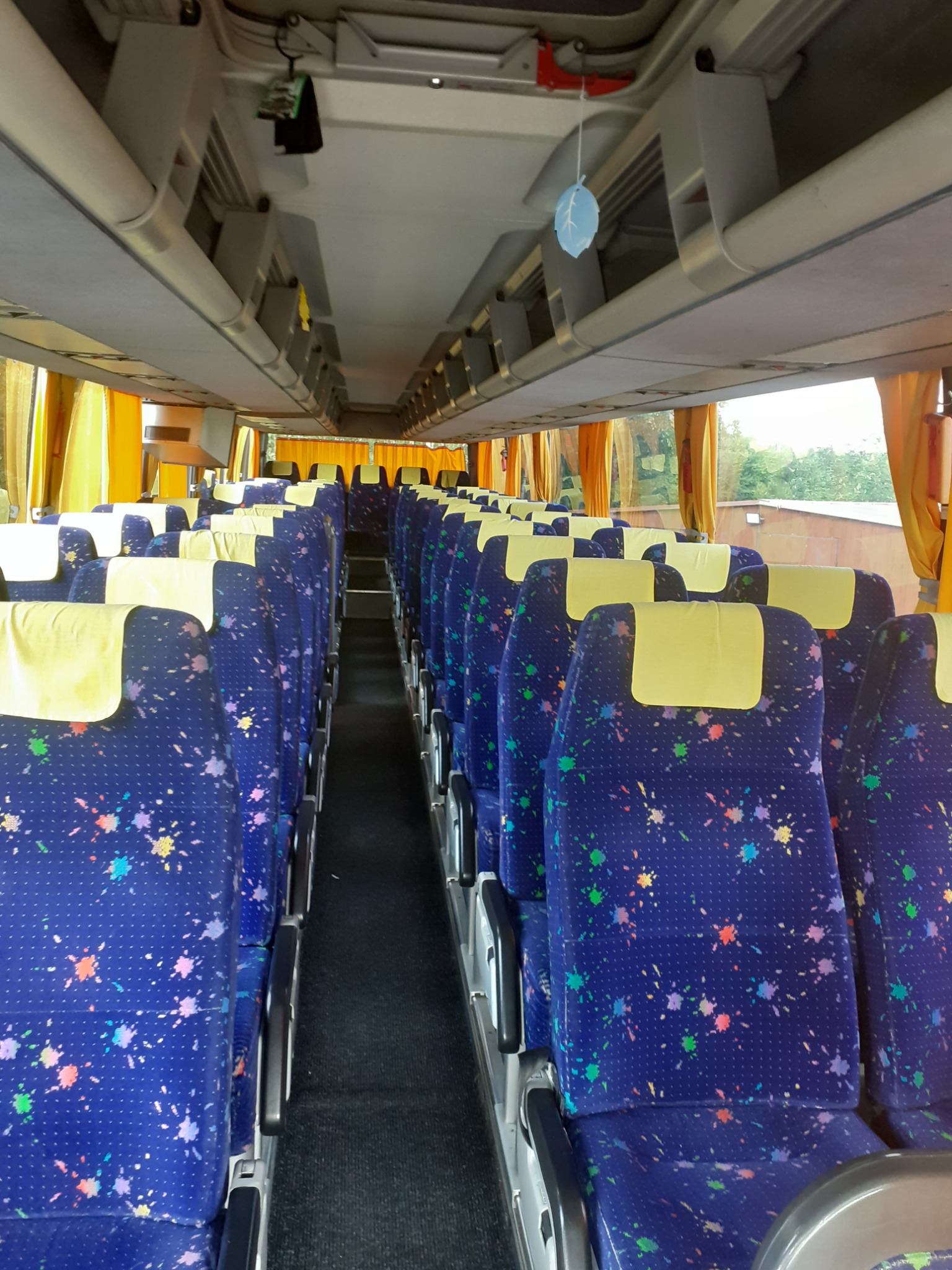 Wnętrze autobusu turystycznego z rzędami niebieskich foteli w kolorowe plamy i żółtymi zagłówkami, widok z przodu na korytarz między siedzeniami, z żółtymi zasłonami w oknach i zawieszką...