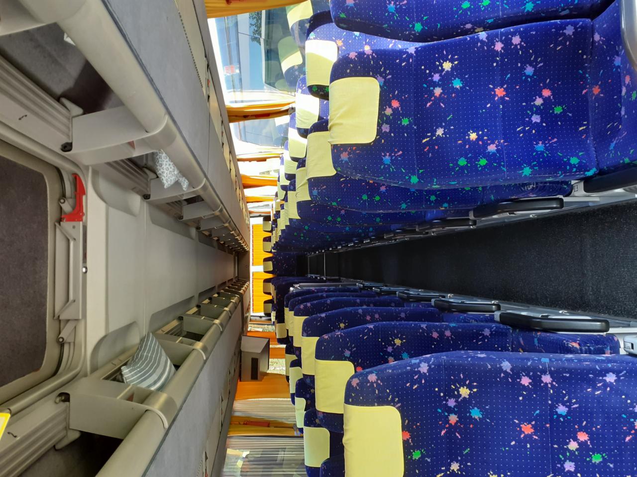 Wnętrze autobusu z rzędami niebieskich foteli z kolorowymi plamkami i żółtymi zagłówkami, widok z góry na dół, z prawej strony półki na bagaż z szarą tapicerką i poduszkami.