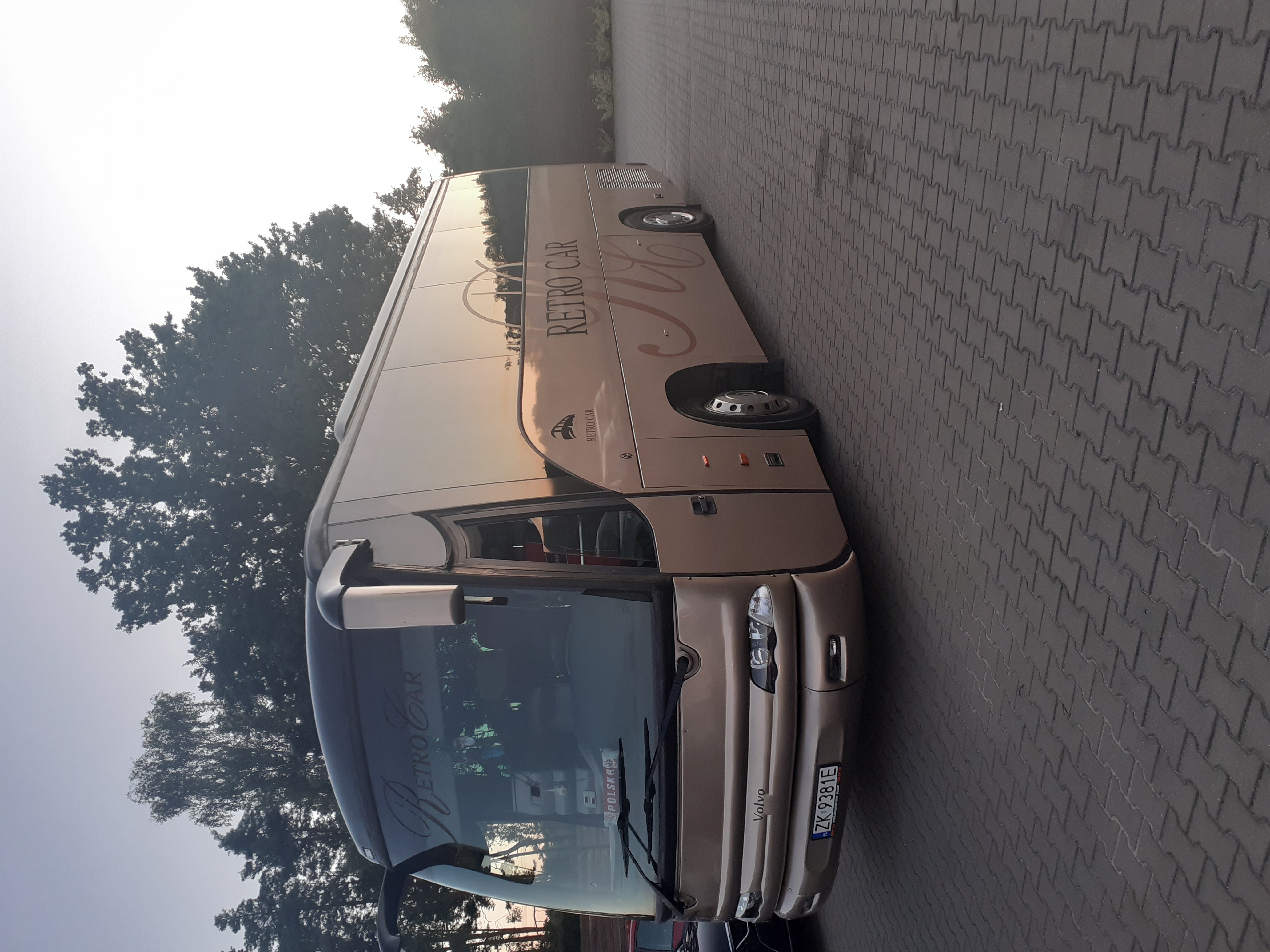 Złoty autokar z napisem 'RETRO CAR' zaparkowany na kostce brukowej, widoczne drzewa w tle.