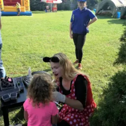 Animator w stroju Myszki Miki maluje twarz dziewczynce na tle dmuchanego zamku i namiotu na zielonej trawie.