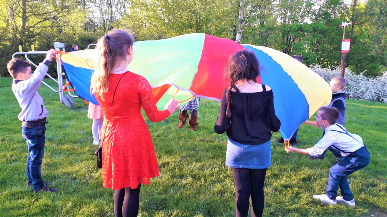 Grupa dzieci bawi się kolorową chustą animacyjną na trawie, w tle drzewa i plac zabaw.
