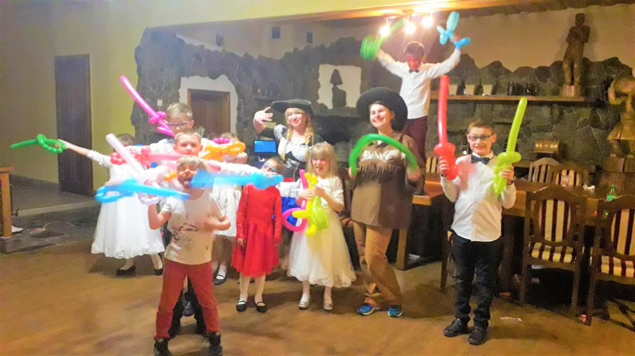 Grupa dzieci i dorosłych w kostiumach, trzymających balony w kształcie mieczy i innych figur, pozujących na tle kamiennej ściany i drewnianych mebli podczas przyjęcia.