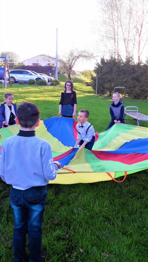 Grupa dzieci bawi się kolorową chustą animacyjną na trawie podczas imprezy plenerowej.