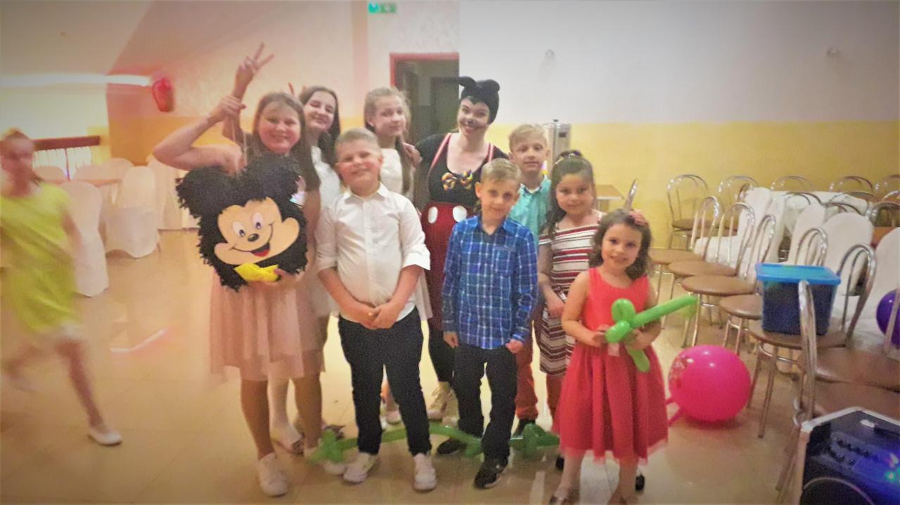 Grupowe zdjęcie dzieci z animatorem przebranym za Myszkę Miki na tle sali z krzesłami, jedna z dziewczynek trzyma balon w kształcie zwierzęcia, a inna maskę Myszki Miki.