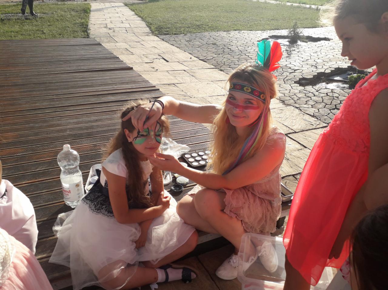 Animator maluje twarz dziewczynki w zielone wzory, obok stoi paleta z farbami, a animator ma na głowie opaskę z piórami i pomalowane oczy na czerwono.