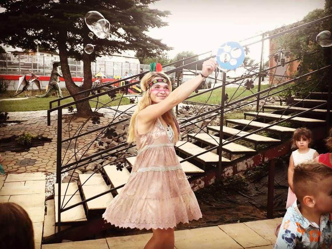 Uśmiechnięta kobieta w stroju inspirowanym indiańskim, z pomalowaną twarzą i pióropuszem, puszcza bańki mydlane, na tle schodów i dzieci w ogrodzie z figurami dinozaurów.