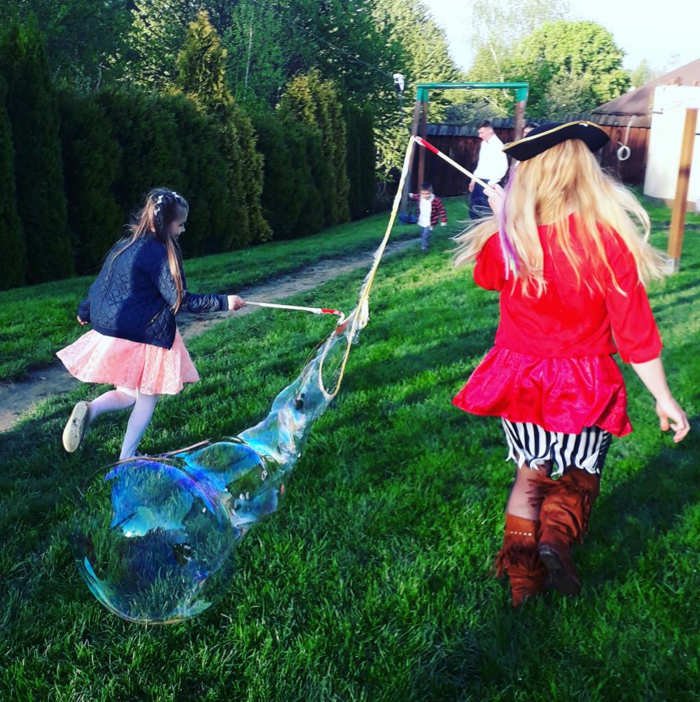 Dwie dziewczynki bawią się na trawie, tworząc ogromne bańki mydlane za pomocą specjalnych kijków; jedna z nich ubrana jest w strój pirata z kapeluszem.