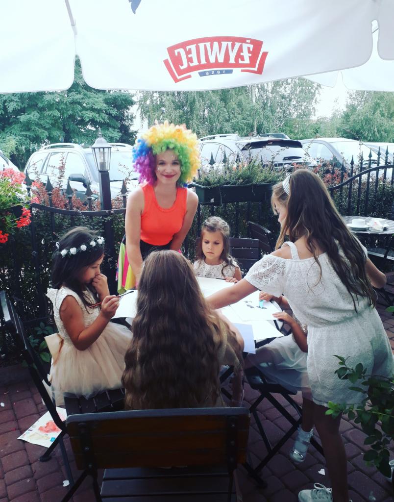 Animator w kolorowej peruce i pomalowaną twarzą zabawia grupę dziewczynek przy stole na zewnątrz, podczas imprezy.