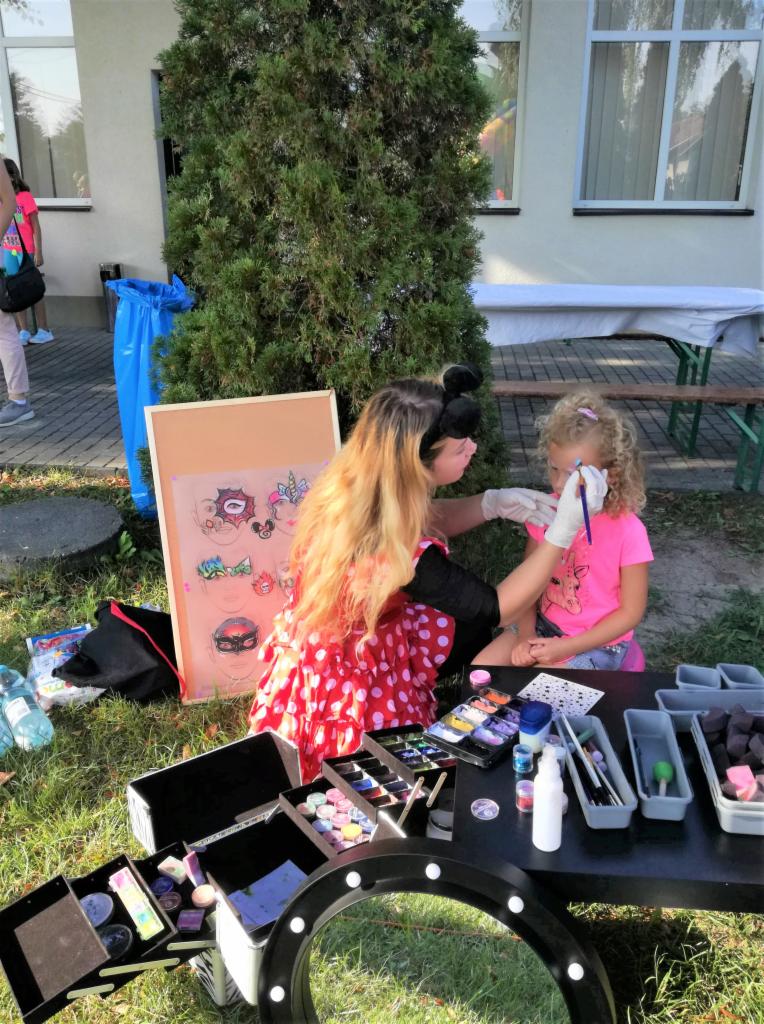 Kobieta w stroju w kropki maluje twarz dziewczynce na trawie, z rozłożonymi kosmetykami i wzornikiem obok.