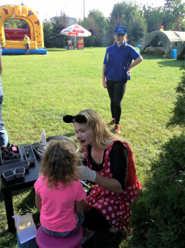 Animator w stroju Myszki Miki maluje twarz dziewczynce na tle dmuchanego zamku i namiotu na zielonej trawie.