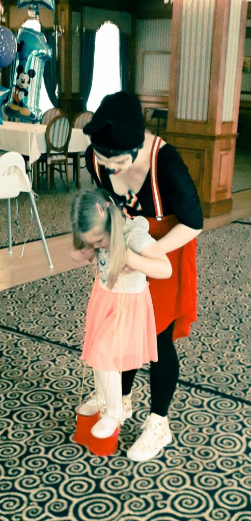 Animator w czarno-czerwonym stroju pomaga dziewczynce w różowej spódniczce chodzić na czerwonych szczudłach w sali z wzorzystym dywanem, w tle dekoracje urodzinowe.