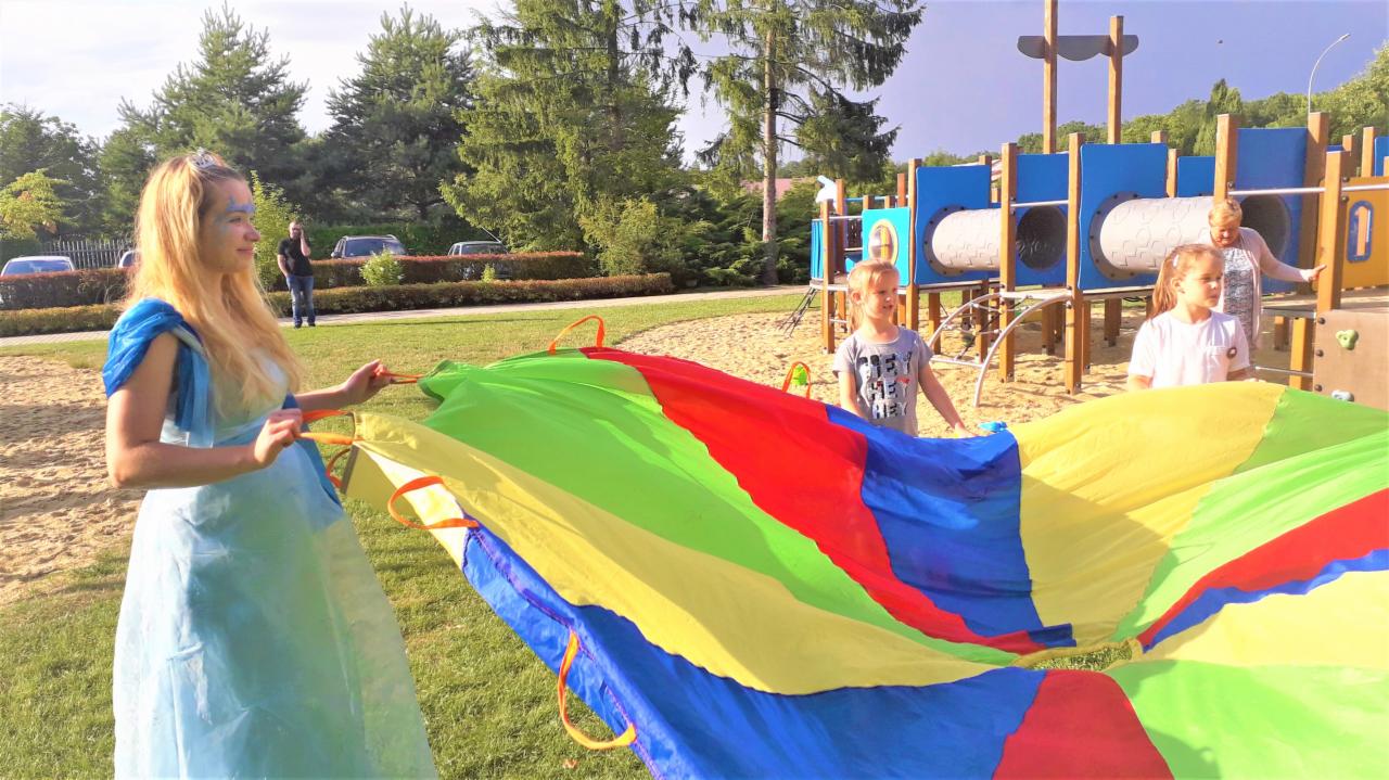 Animator w niebieskiej sukience i z pomalowaną twarzą trzyma kolorową chustę animacyjną z dziećmi na tle placu zabaw.