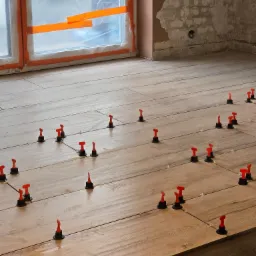 Ułożone płytki podłogowe imitujące drewno, ze specjalnymi czerwono-czarnymi klipsami poziomującymi i krzyżykami dystansowymi, okno zabezpieczone folią malarską, surowa ściana w tle.