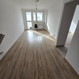 Uslugi Krzysztof Szczepaniak - Jasne wnętrze po remoncie: parkiet w odcieniach brązu, białe ściany, okno z widokiem na balkon. Minimalistyczne krzesło i wiszące kable dodają surowego charakteru.