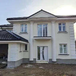 Elewacja dwukondygnacyjnego domu z kolumnami w stylu klasycystycznym, z garażem w trakcie wykańczania, widoczny samochód dostawczy z reklamą firmy budowlanej.