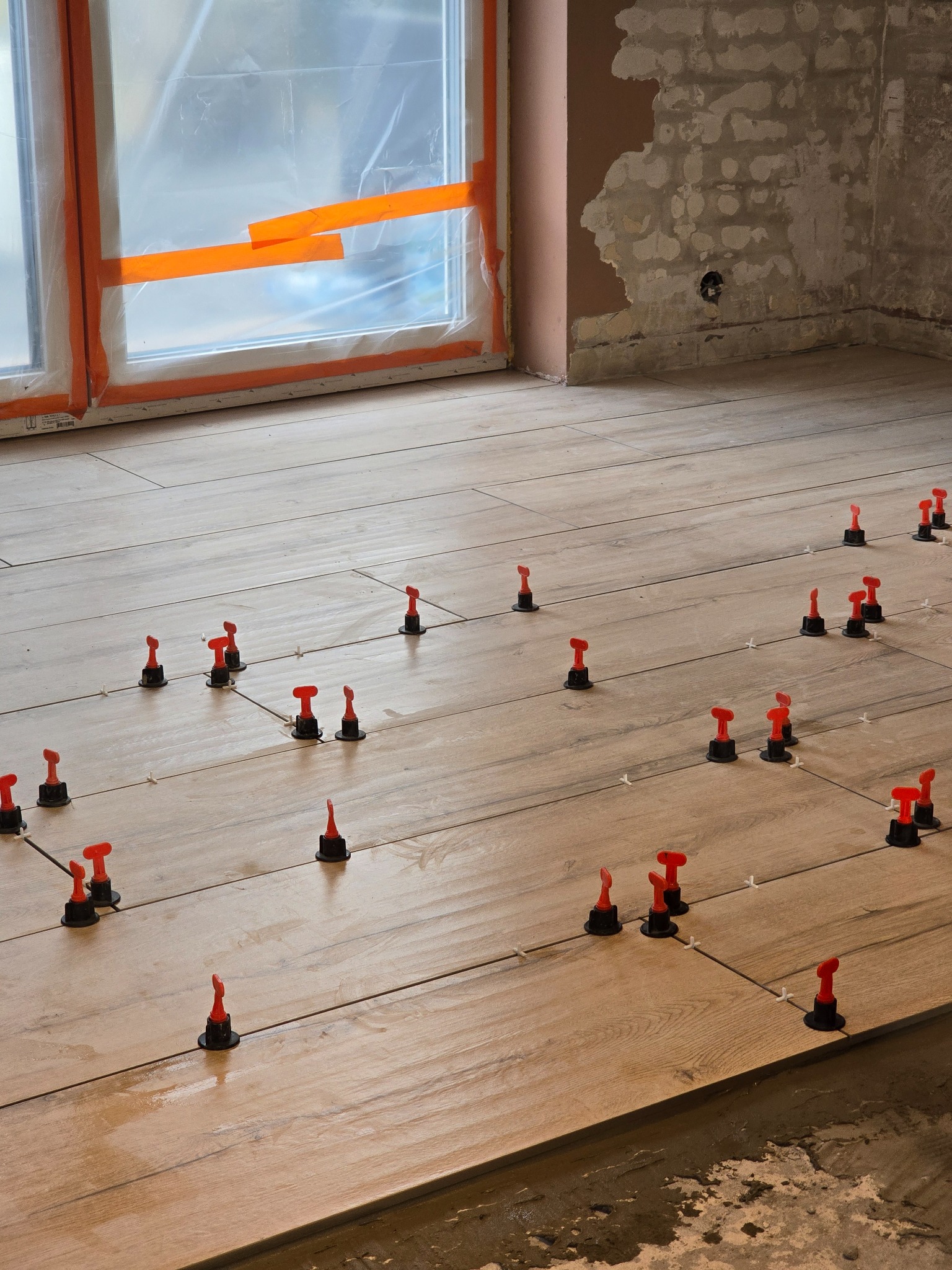 Ułożone płytki podłogowe imitujące drewno, ze specjalnymi czerwono-czarnymi klipsami poziomującymi i krzyżykami dystansowymi, okno zabezpieczone folią malarską, surowa ściana w tle.