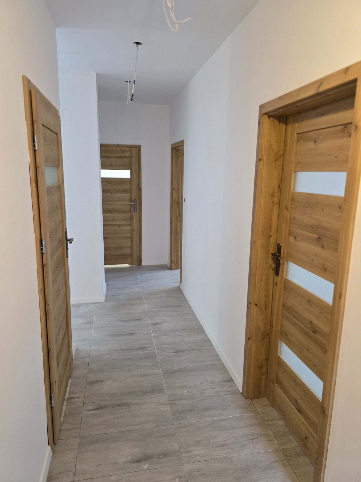 Korytarz z szarymi płytkami podłogowymi imitującymi drewno, białymi ścianami i trzema drewnianymi drzwiami z przeszkleniami, widoczne niedokończone podłączenie oświetlenia sufitowego.