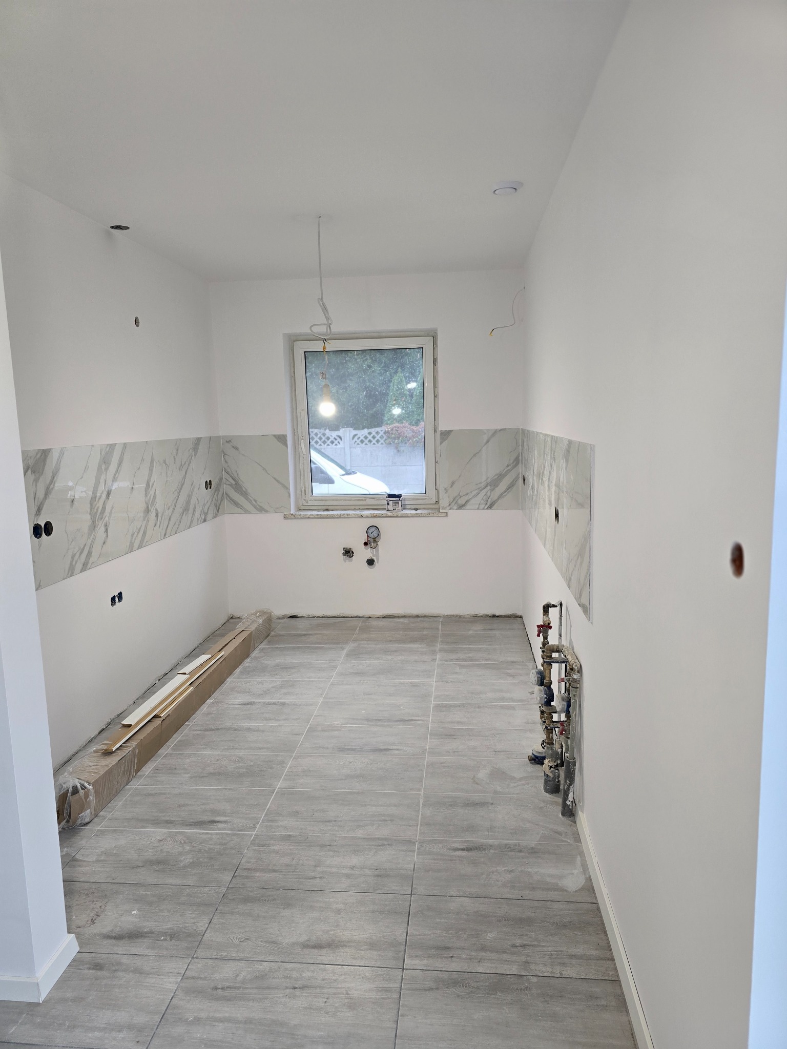 Wnętrze kuchni w trakcie remontu: położone szare płytki imitujące drewno na podłodze, częściowo wykończone ściany z białymi płytkami z marmurowym wzorem, widoczne instalacje wodne i elektryczne...