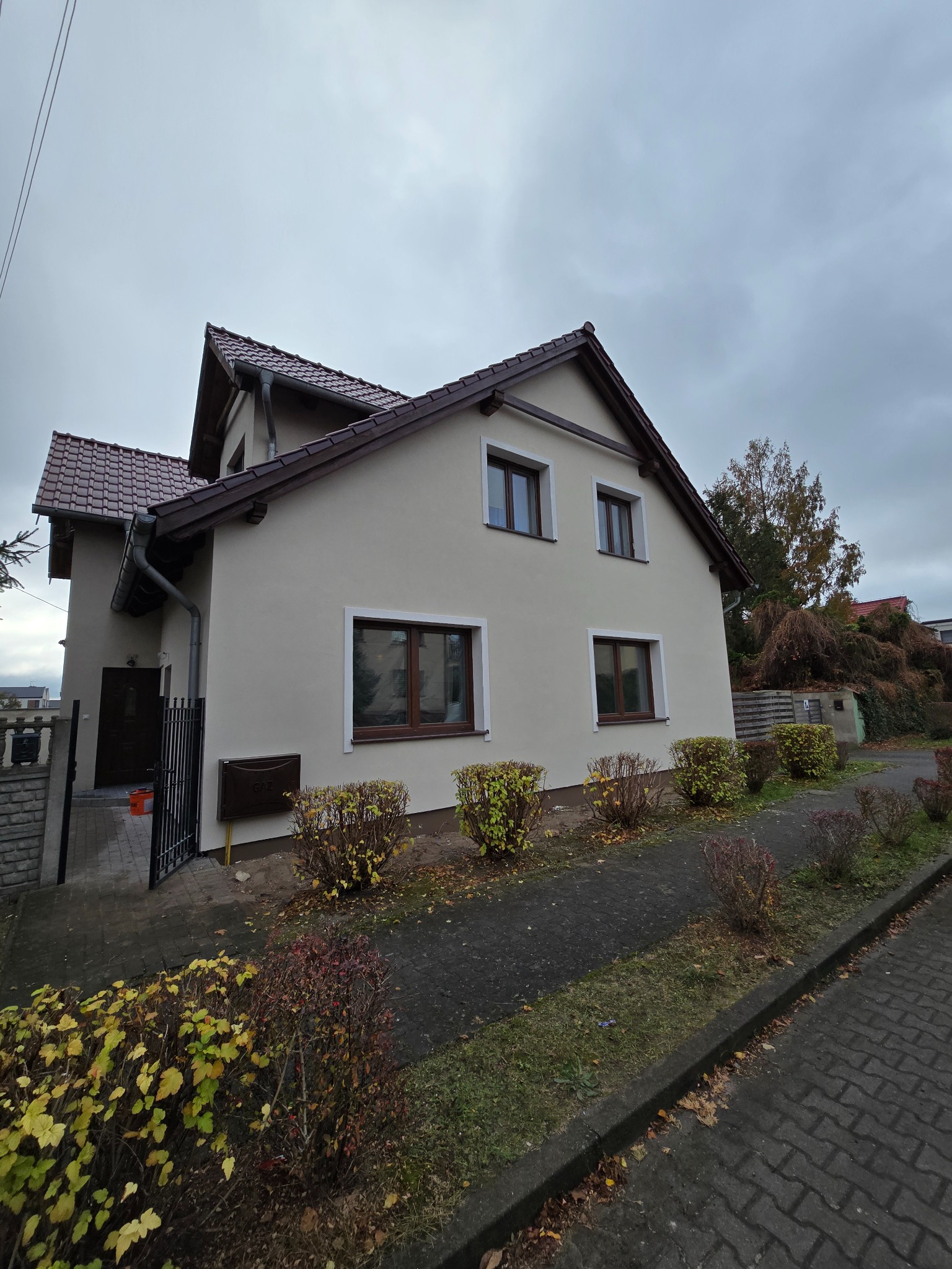 Dwupiętrowy dom jednorodzinny z jasnobeżową elewacją, brązowymi ramami okiennymi i ciemnobrązowym dachem z dachówki, sfotografowany w pochmurny dzień z chodnika obok niskich krzewów.