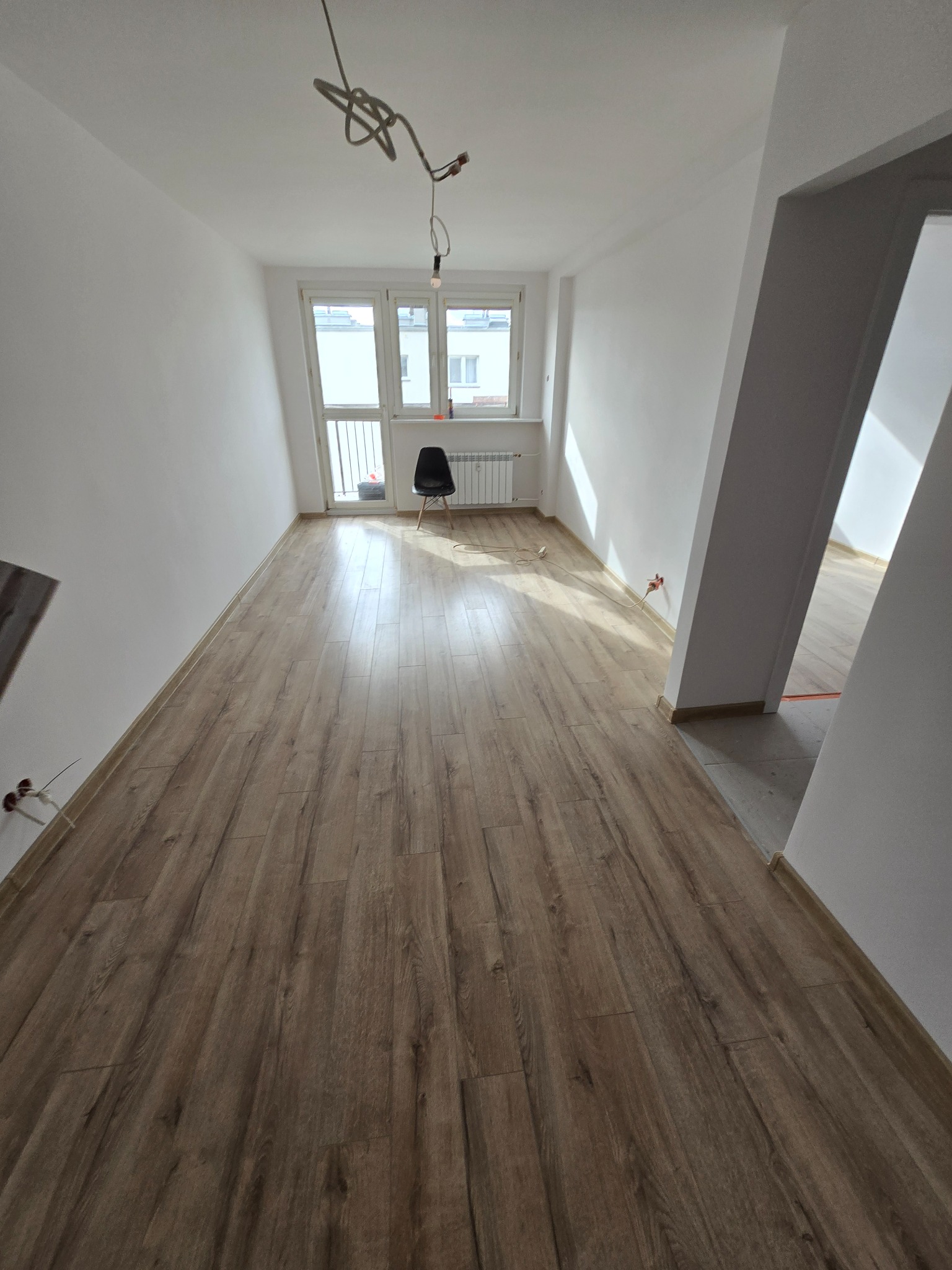 Jasne wnętrze po remoncie: parkiet w odcieniach brązu, białe ściany, okno z widokiem na balkon. Minimalistyczne krzesło i wiszące kable dodają surowego charakteru.