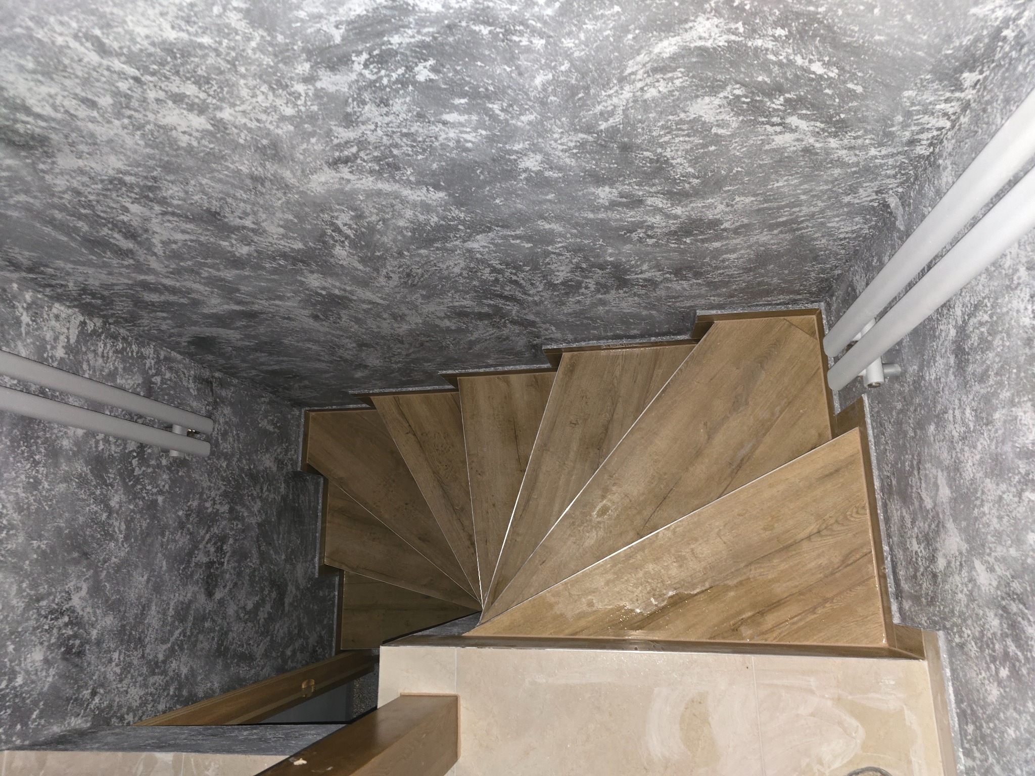 Spiralne schody z drewnopodobnych płytek, widok z góry. Dekoracyjna szara ściana i sufit z efektem betonu. Białe rury grzewcze wzdłuż ściany.