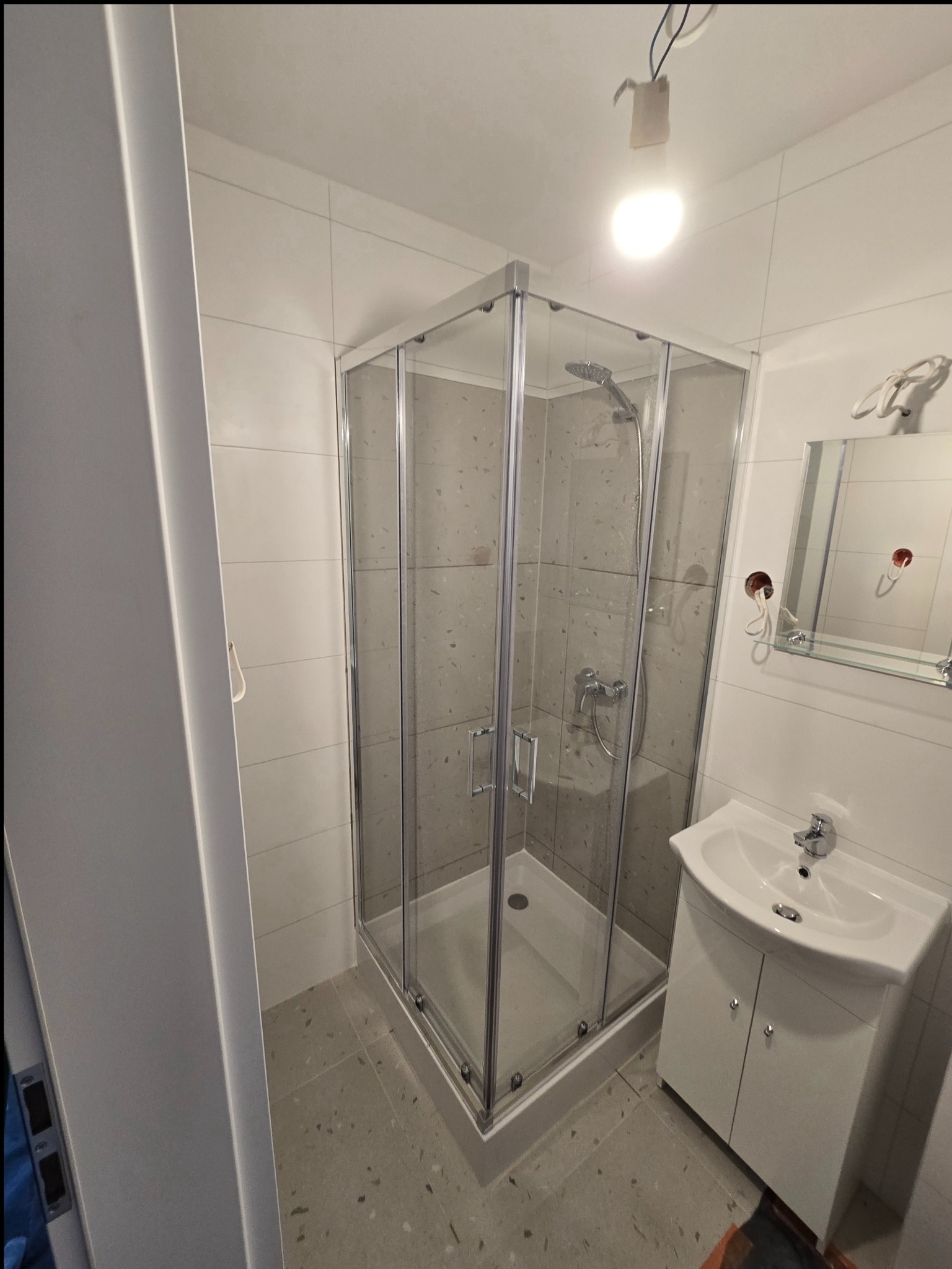 Nowoczesna kabina prysznicowa z brodzikiem, umywalka z szafką i lustro w wykończonej łazience. Jasne płytki i szara podłoga z terazzo.