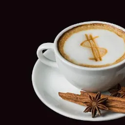 Biała filiżanka kawy z symbolem dolara zrobionym ze spienionego mleka, leżąca na białym spodku z laskami cynamonu i anyżem gwiazdkowym na czarnym tle.