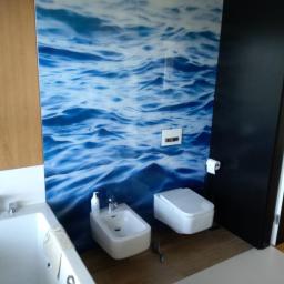 Mateusz Bobrowski - Nowoczesna łazienka z białą armaturą (wanna, bidet, toaleta) i fototapetą z motywem fal morskich, widoczna część szafki z drewnianymi frontami oraz czarne drzwi.