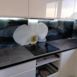 Szklany panel kuchenny z motywem białej orchidei na tle kamieni, podświetlony listwą LED, zamontowany nad płytą indukcyjną w nowoczesnej kuchni z białymi szafkami.