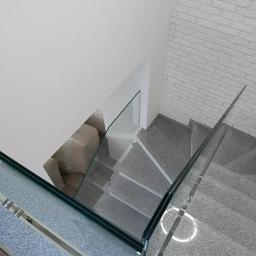Mateusz Bobrowski - Schody wykończone szarym kamieniem z balustradą ze szkła hartowanego, widok z góry, ściana z białej cegły w tle.