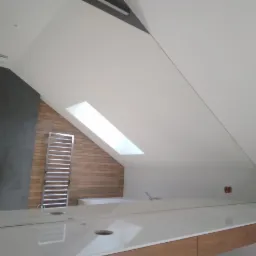 Nowoczesna łazienka na poddaszu z oknem dachowym, widoczna umywalka z blatem z jasnego marmuru i drewniane szafki pod spodem, grzejnik drabinkowy na ścianie z drewnianą okładziną. Nietypowy układ...