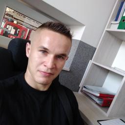 Mateusz Bobrowski - Mężczyzna w czarnej koszulce siedzi na krześle; w tle widać wóz strażacki za oknem i regał z segregatorami.