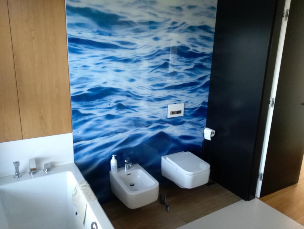 Nowoczesna łazienka z białą armaturą (wanna, bidet, toaleta) i fototapetą z motywem fal morskich, widoczna część szafki z drewnianymi frontami oraz czarne drzwi.