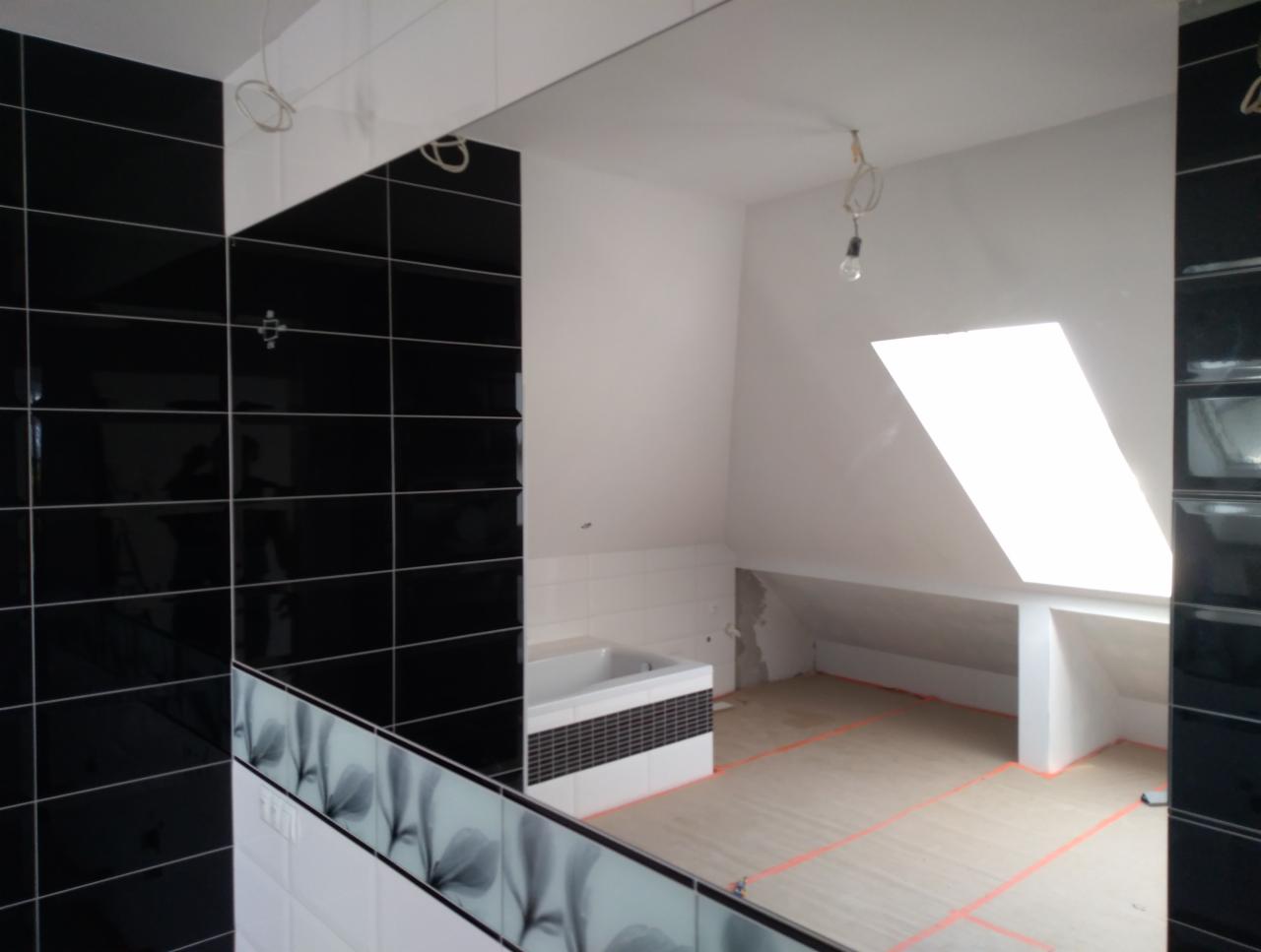 Częściowo wykończona łazienka na poddaszu z czarno-białymi płytkami i widocznym oknem dachowym, odzwierciedlona w dużym lustrze, z widocznymi przewodami elektrycznymi i przygotowanym podłożem...