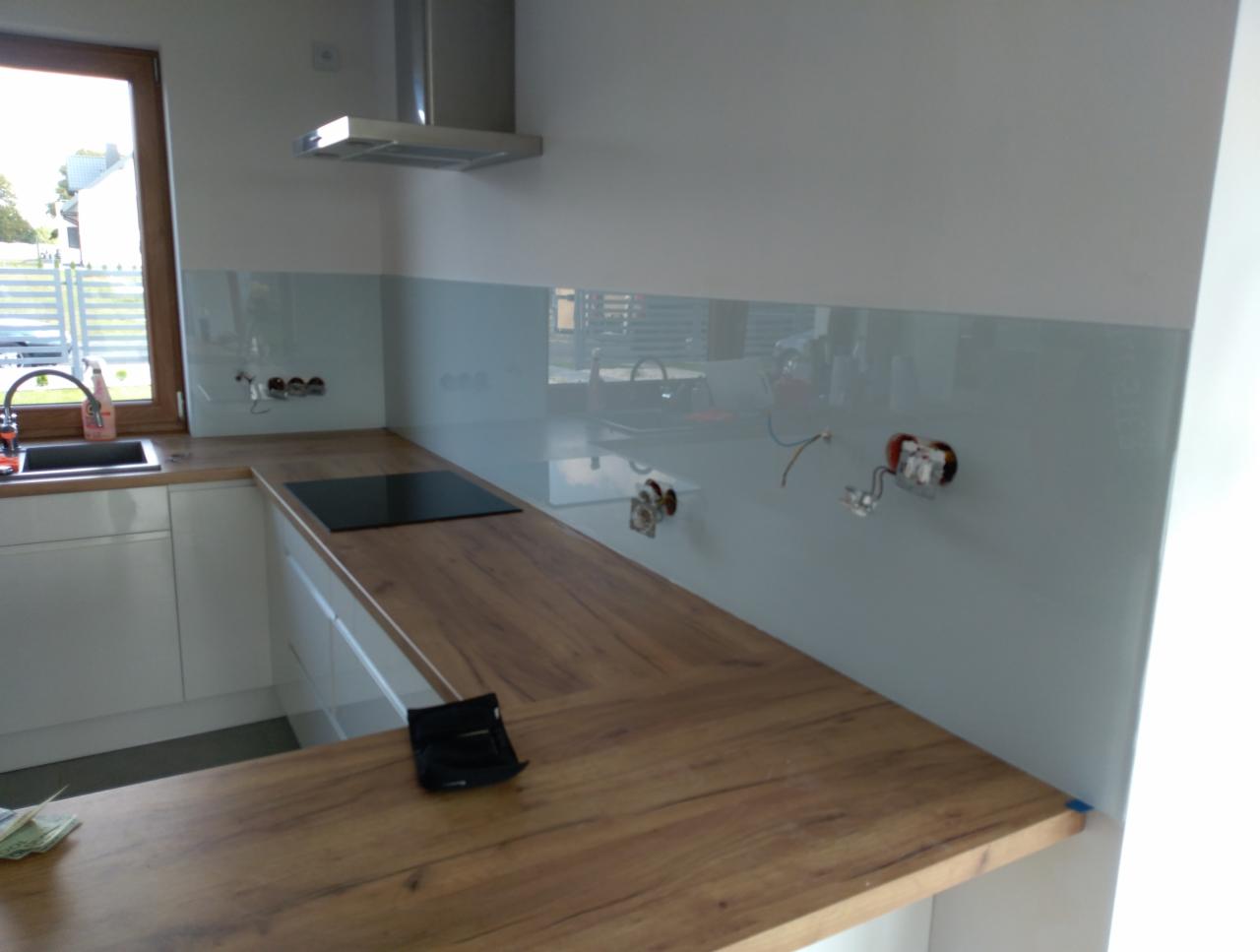 Nowoczesna kuchnia z drewnianym blatem, białymi szafkami i szklanym panelem ściennym w odcieniach szarości, z widocznymi puszkami elektrycznymi przed podłączeniem.